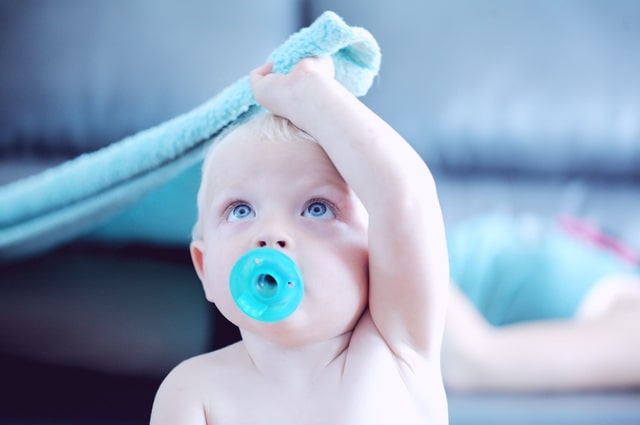 Most Popular Baby Boy Names Right Now, a picture of a baby boy, sucking on a blue pacifier and pulling on a blue baby blanket.